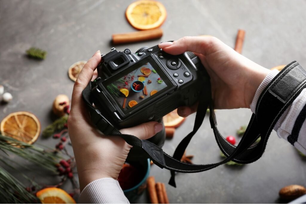 Mani che tengono una fotocamera reflex con display acceso mentre inquadra una composizione creativa di elementi natalizi e spezie. Scena ambientata su sfondo scuro con styling visivo curato. dmplus. dm+. Damiano Macaluso