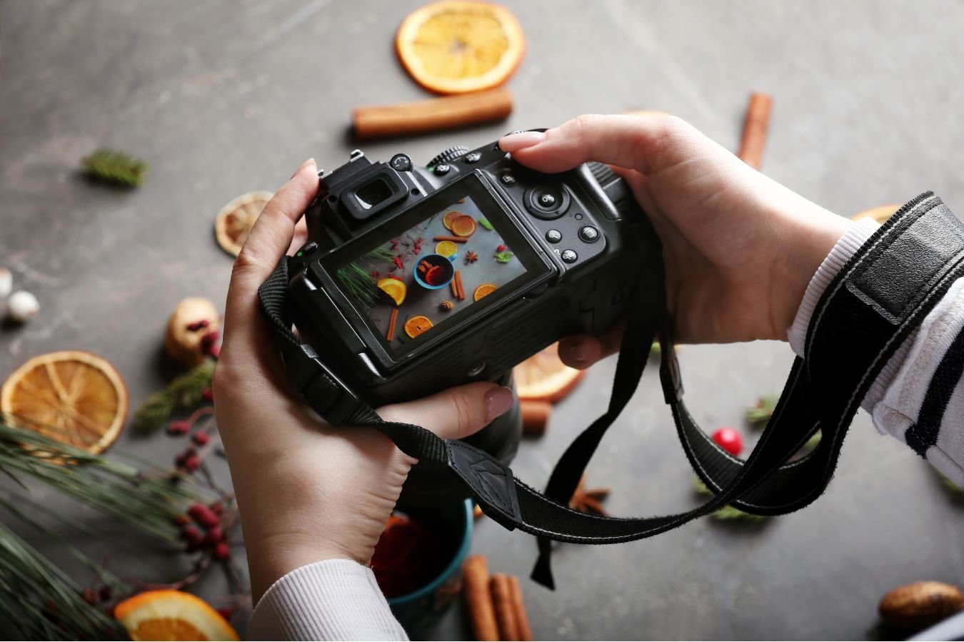 Mani che tengono una fotocamera reflex con display acceso mentre inquadra una composizione creativa di elementi natalizi e spezie. Scena ambientata su sfondo scuro con styling visivo curato. dmplus. dm+. Damiano Macaluso