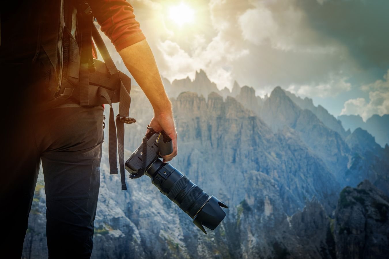 Fotografo in montagna con zaino e fotocamera in mano, ripreso di spalle mentre osserva un paesaggio roccioso al tramonto. dm+. Damiano Macaluso